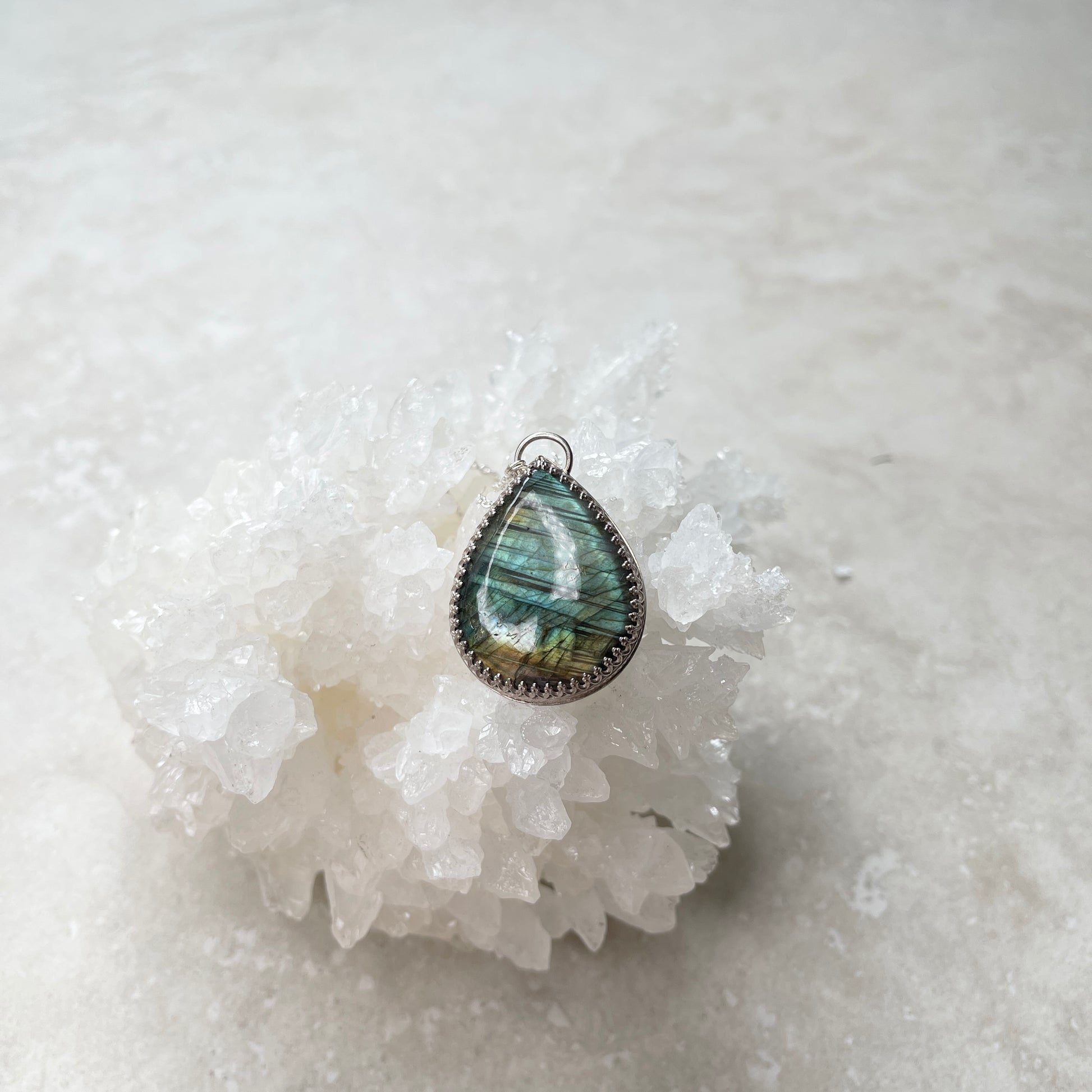 A Labradorite pendant necklace on a cable chain, with the pendant resting on a white crystal. The necklace features a teardrop-shaped stone with a metallic silver frame.